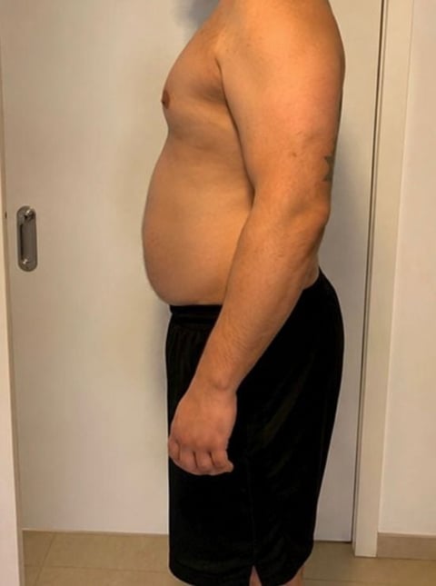 Man with prominent belly standing shirtless in black shorts against white wall